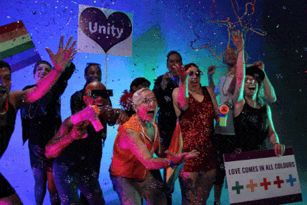 CP24_Pride-Toronto_PhotoboothTO-3D-Camera-Array A group of people at a pride event celebrate under colorful lights, holding rainbow flags and signs reading “Unity” and “Love Comes In All Colours.” Confetti falls around them.