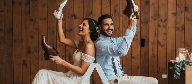 Bride and groom sit back-to-back holding up each other's shoes, smiling, during a wedding game in front of a wooden wall.